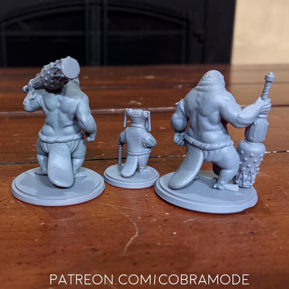 three figurines of fantastical creatures, including a large, muscular creature with a large head and a smaller creature with a long tail. The figurines are displayed on a wooden surface, and there is text in the image that reads "Patron.comi cobramode".